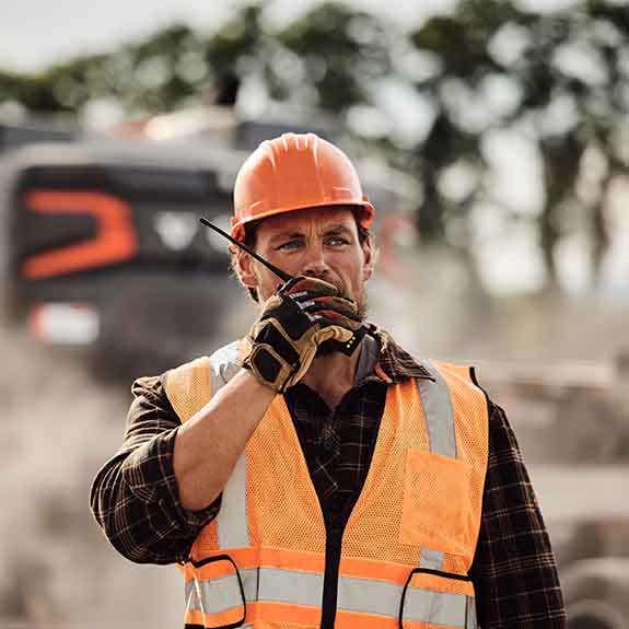 A construction worker speaking on a walkie-talkie with the Volvo VNX in the background.