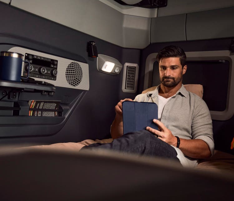 A man ralzing in the spacious seating reading something on his tablet with the light on. Unwinding in the Volvo VNX after a long day.