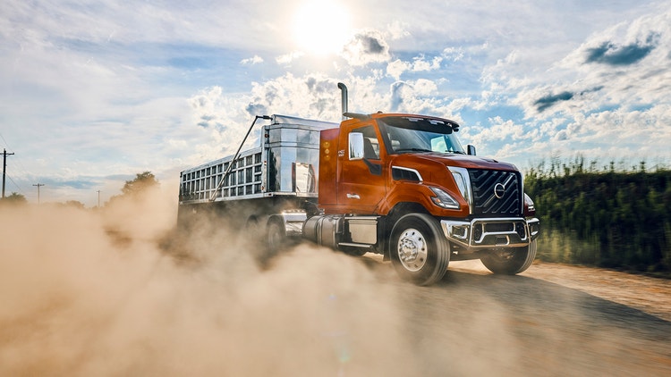A reddish orange color Volvo VNX truck with a dusty smoke around the wheels of the truck.
