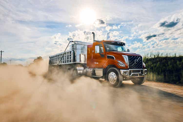 An orange colored Volvo VNX on a muddy road with dusty smoke aurrounding the wheels of the truck.